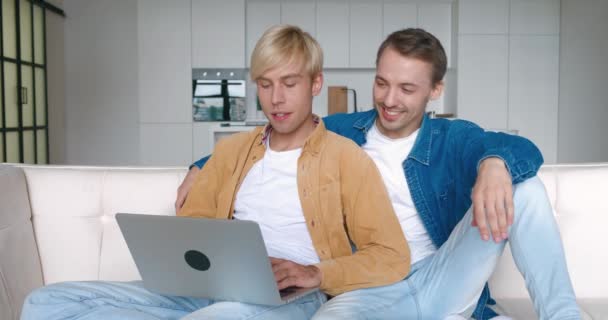 Dos hombres gay pareja usando ordenador portátil para videollamadas, hablando distantemente con amigos. Dos hombres LGBTQ teniendo video confetencia mientras descansan en el sofá en casa — Vídeo de stock