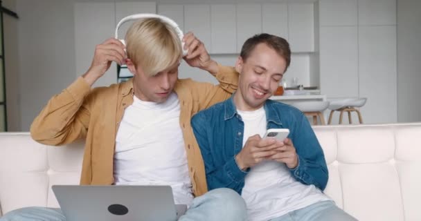 A férfi meleg pár otthon tölti az idejét. Két LMBTQ férfi pihen a kanapén és zenét hallgat okostelefonnal és fejhallgatóval a kanapén a nappaliban — Stock videók