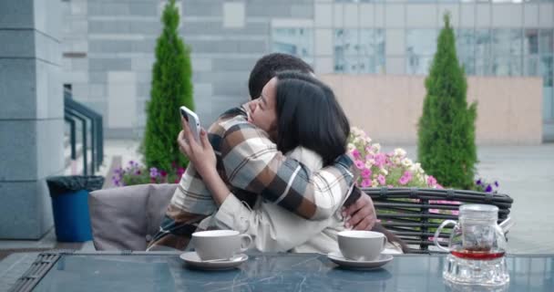 Two charming people in casual cozy clothes sitting at the table in a summer terrace of a cafeteria on sunny warm early spring day and looking at a mobile phone, rejoicing and hugging each other. — 图库视频影像