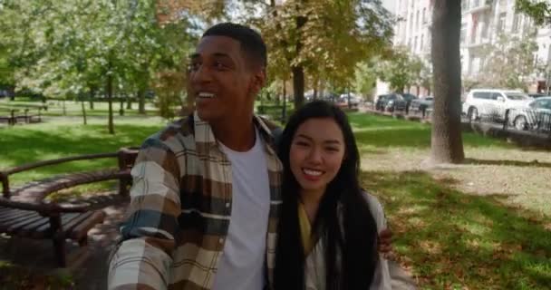 Happy multiracial young loving couple actively walking in the park and recording video or having video call on mobile phone in the outstretched of a handsome African guy hugging his pretty girlfriend — 图库视频影像