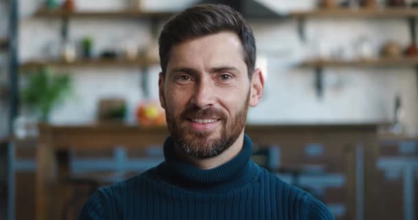 Retrato de cerca del exitoso hombre caucásico sonriendo mirando a la cámara mientras está sentado en casa en el sofá. Cabeza de hombre barbudo sobre fondo de cocina — Vídeos de Stock