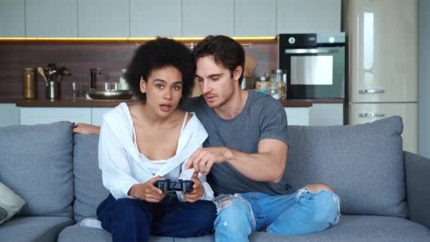 Olika pojkvän och flickvän sitter tillsammans på soffan i vardagsrummet och klicka på joystick medan aktivt spela på dataspel, titta på skärmen. — Stockvideo