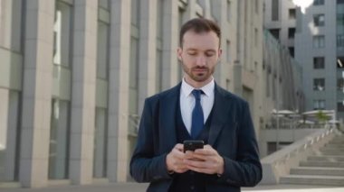 Modern ofis binasının yakınında akıllı telefon kullanan dalgın bir erkek işadamı. Klasik takım elbiseli şık sakallı adam cep telefonuna mesaj yazıyor. Sosyal ağ. Uygulamalar. Akıllı telefonlar.