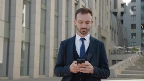 관대 한 남성 사업가가 스마트폰을 사용하여 현대 사무실 건물 근처 시내에 서 있습니다. 클래식 수트를 입고 휴대폰으로 메시지를 입력하는 세련 된 수염의 남자. 소셜 네트워크. 앱. 스마트폰. — 비디오