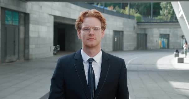 Portrait handsome businessman in glasses walking outside near office building. Redheaded man in formal suit looking away outdoors at city — стоковое видео
