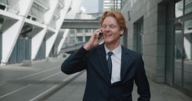 Resmi takım elbiseli gülümseyen yakışıklı iş adamı şehir merkezindeki ofis binalarının yanında beklerken duygusal olarak cep telefonuyla konuşuyor. Başarılı bir iş adamı iyi bir anlaşma yapıyor.