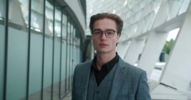 Gülümseyen CEO yöneticisinin yakın plan portresi gözlük takıp kameraya bakıyor. Modern ofis binasının yanında duran, gözlüklü, klasik takım elbiseli, kendinden emin bir erkek işadamı.