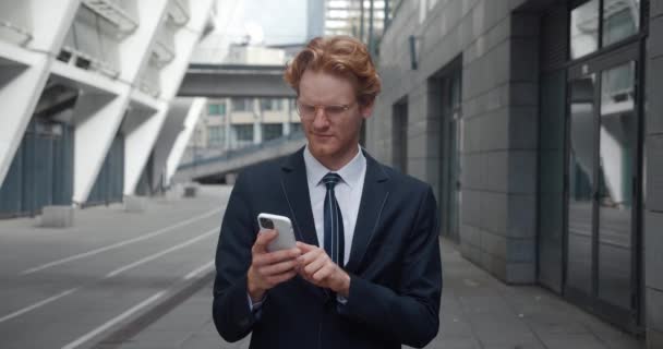 Стильний рудий бізнесмен, використовуючи смартфон, що стоїть на вулиці на вулиці ділового району в центрі міста. Успішний чоловічий підприємець використовує мобільний додаток електронної комерції для інвестицій на фондовому ринку — стокове відео