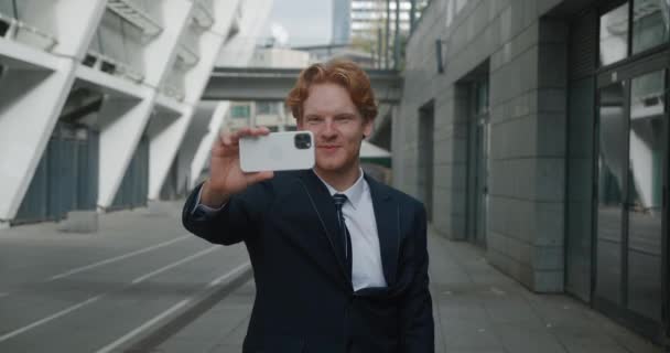 Lachende knappe roodharige zakenman die selfie video chat met behulp van smartphone buiten in de stad straat. Mannelijke ondernemer in formele pak praten videogesprek op mobiele telefoon in het centrum district. — Stockvideo