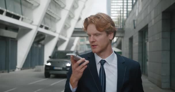 Concentrated businessman recording voice message standing outdoors at business district. Emotional red-haired male entrepreneur talking on mobile phone speakerphone, conducting aggressive negotiations — Stock Video