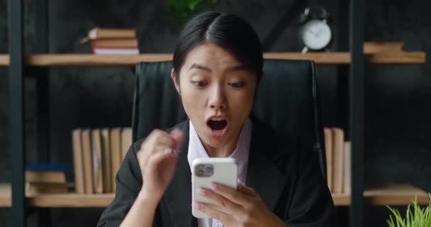 幸せなビジネス女性の肖像は、現代のオフィスに座ってスマートフォンで成功をお楽しみください。屋内で携帯電話で勝利を祝う女性 — ストック動画