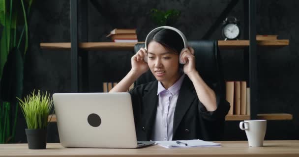 Gelukkig aziatische zakenvrouw is tevreden met het werk gedaan op de laptop computer, zet op koptelefoon en het luisteren van muziek, genieten van succes in het bedrijfsleven tijdens het zitten in modern kantoor werkplek. — Stockvideo