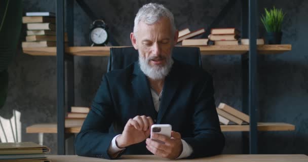 Smiling mature bearded businessman uses smartphone while sitting in office. Stylish entrepreneur boss uses a mobile phone at his workplace. — Wideo stockowe