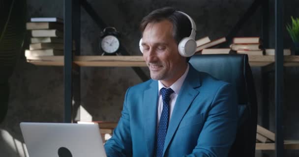 Portrait of smiling mature male businessman enjoying music with headphones while working with laptop at modern office. — Stock Video