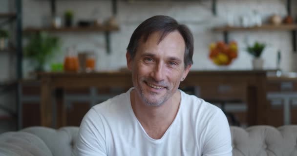 Close-up portrait of smiling caucasian mature man looking at camera while sitting at home on sofa. Headshot of happy male on kitchen background — ストック動画