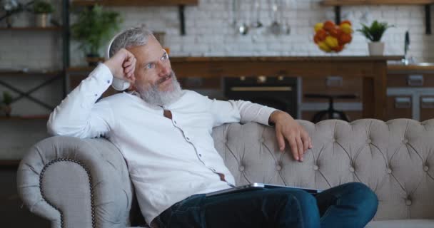 Potret pria berjanggut dewasa dengan rambut abu-abu menikmati musik dengan headphone nirkabel dan menggunakan komputer laptop saat beristirahat di sofa di apartemen ruang tamu — Stok Video