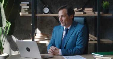 Olgun erkek iş adamı CEO ofis masasında otururken dizüstü bilgisayarla video görüşmesi yapıyor. İş elbiseli bir adam web kamerasıyla uzaktan görüşüyor. Teknoloji ve iletişim