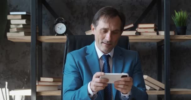 Glimlachende volwassen zakenman spelen op zijn smartphone spannende mobiele applicatie winnende racespel knijpen vuist vreugde tijdens het zitten op modern kantoor — Stockvideo