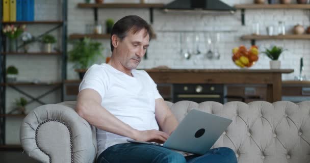 シニア成熟した男タイプのラップトップブラウジングインターネット上のアプリケーションを使用して座っているソファだけで自宅アパートで — ストック動画