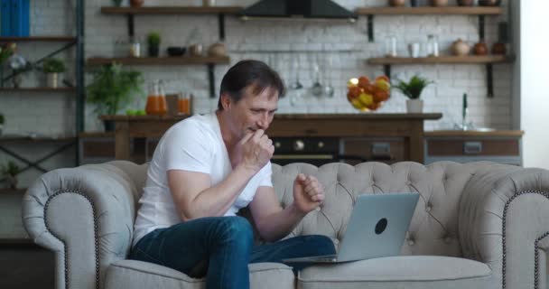 Onaangenaam verrast en geschokt volwassen man door het slechte nieuws te zien op het laptopscherm tijdens het werken op de bank thuis. Geschokte man lijdt onder stress en wanhoop — Stockvideo