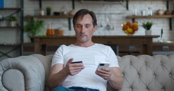 Happy mature man sitting on sofa at living room, holding credit card and mobile phone to make online transaction, buying wished items, enjoying shopping from home concept — ストック動画