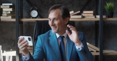 Modern ofisteki akıllı telefonuyla görüntülü konuşma yapan mutlu bir iş adamı telefon ekranında insanları selamlıyor. Takım elbiseli erkek patron iş yerinde cep telefonuyla selfie görüntülü konuşma yapıyor.