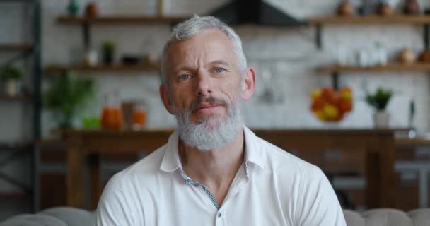 Cheerful bearded man with grey hair sitting on couch, looking at camera. — Stockvideo