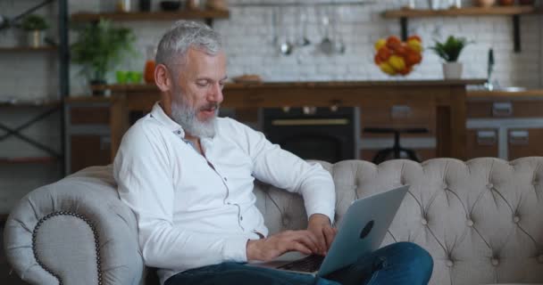 Retrato de alegre caucásico guapo hombre barbudo maduro con pelo gris sentado en el sofá en la sala de estar en el apartamento y escribiendo en el ordenador portátil, trabajando a distancia en casa — Vídeo de stock