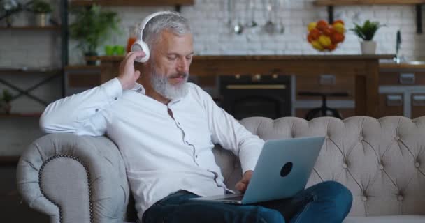 Portrait of mature bearded man with grey hair enjoying listening music with wireless headphones and using laptop computer while sitting on sofa in living room apartment — Stock videók