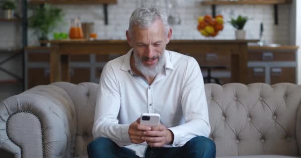 Amazed happy mature businessman receiving message reading good news at living room. Excited overjoyed male winner celebrating success looking at smart phone on sofa at home. Mobile victory concept — Stockvideo