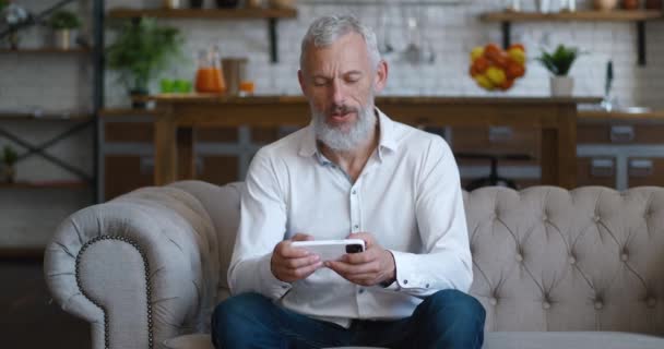 Besorgter bärtiger älterer Mann spielt online Videospiel mit Smartphone-App, sitzt zu Hause auf dem Sofa, enttäuscht über den Verlust einer Online-Spielanwendung — Stockvideo