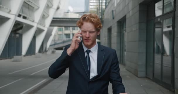 Hombre de negocios guapo de confianza en traje formal hablando emocionalmente por teléfono móvil mientras está parado afuera en el centro cerca de edificios de oficinas. Hombre de negocios exitoso negociando un buen trato — Vídeos de Stock