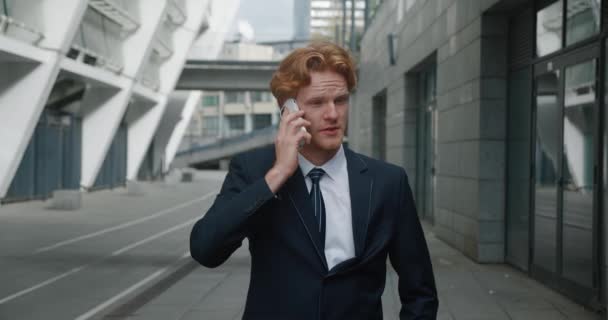 Confident handsome businessman in formal suit emotionally talking by mobile phone while standing outside at downtown near office buildings. Successful businessman negotiating a good deal — Wideo stockowe