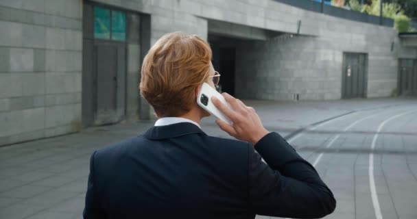 Zadní pohled na sebevědomého mladého podnikatele ve formálním obleku mluvící telefon při procházce v centru v obchodní čtvrti. Usmívající se rusovlasý muž-podnikatel mluví na mobilním telefonu, zatímco jde do práce. — Stock video