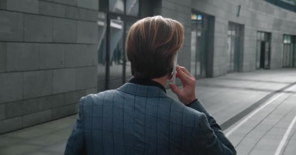 Achteraanzicht van ambitieuze zakenman praten op mobiele telefoon en lopen door het zakendistrict. Achteraanzicht van zelfverzekerde jonge mannelijke ondernemer communiceren aan de telefoon op weg naar zijn werk. — Stockvideo