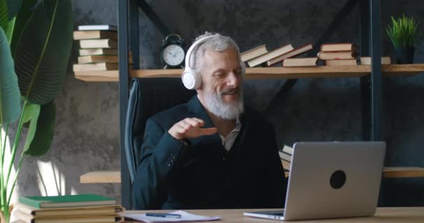 Joyful mature business man dancing in workplace wearing wireless headphones listening music while working at laptop computer sitting at office — Vídeo de Stock