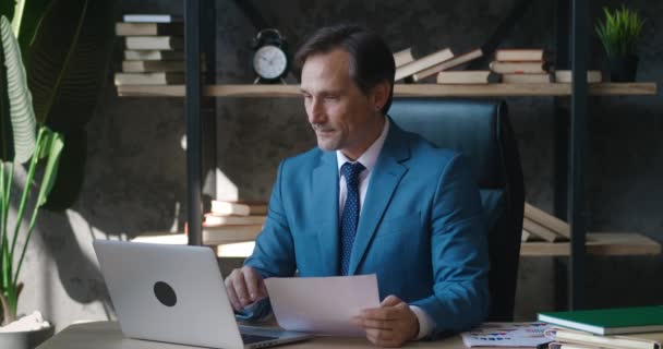 Mature busy business man working at laptop computer and with paper documents analyzing sales charts while sitting at table at modern office. — Stock Video