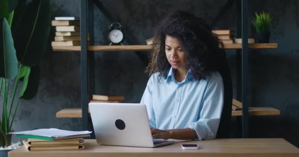 Afro-Amerikaanse jonge vrouw met krullend haar werkend op laptop, kijkend naar smartphone op kantoor. Vrouwelijke student typen op computer toetsenbord binnen. Zakelijke vrouw op zoek naar laptop scherm binnen — Stockvideo