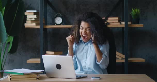 Emocionada joven mujer de negocios afroamericana disfrutando de ganar se sienta a la mesa con el ordenador portátil trabajando en la oficina moderna. Ella recibe grandes noticias y está encantada. Mostrando sí gesto — Vídeo de stock