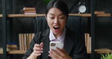Heyecanlı Asyalı iş kadını akıllı telefonu elinde tutarak neşeli bayan kazananı kutluyor online teklif oyunu kazanıyor zafer kazanıyor modern ofiste otururken cep telefonundan iyi haberler okunuyor