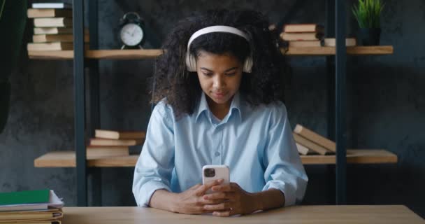 Cheerful african american business woman in headphones holding smartphone texting message and listening music in office. Young female student using mobile phone technology business app — Wideo stockowe
