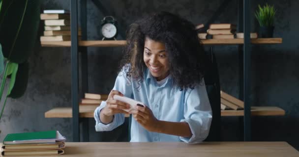 Mujer afroamericana positiva concentrada joven usando teléfono inteligente sentado en el escritorio de la oficina, jugando juegos de carreras en línea aplicación por teléfono móvil hacer sí gesto ganador — Vídeo de stock