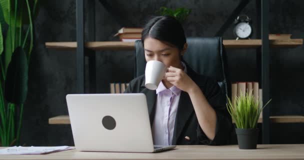 Young asian attractive female office worker sitting at the laptop computer at the desk, working and drinking coffee. Focused manager businesswoman typing on computer keyboard indoors in modern office — ストック動画