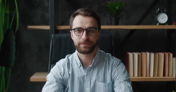 Retrato de hombre de negocios profesional barbudo en gafas girando y mirando a la cámara y sonriendo mientras está sentado en la oficina moderna. — Vídeo de stock