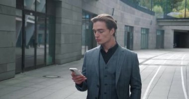 Genç beyaz erkek işadamı modern iş binasında yürürken akıllı telefonuyla yazı yazıyor. Resmi takım elbiseli kendine güvenen yakışıklı adam işe giderken sosyal ağları cep telefonundan kontrol ediyor.