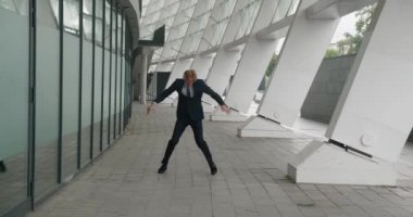 Kablosuz kulaklıklı, müzik dinleyen ve ofis binasının dışında dans eden mutlu bir iş adamı. Resmi takım elbiseli, pozitif kızıl saçlı erkek girişimci müzikten zevk alıyor, başarılı anlaşmayı kutluyor..