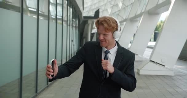 Cheerful red-haired businessman wearing wireless headphones enjoying listening to music with smartphone and dances funny, standing outdoors near an office building — стоковое видео