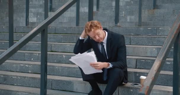 Tired red haired businessman with business papers in his hands sits on the steps of an office building. Sad overworked man employee, putting hand to the head. The concept of workload and burnout. — стоковое видео