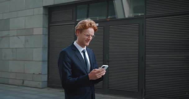 Νεαρός καυκάσιος επιχειρηματίας δακτυλογραφεί σε smartphone, ενώ το περπάτημα κατά μήκος σύγχρονο επιχειρηματικό κτίριο. Χαμογελώντας κοκκινομάλλης με επίσημο κοστούμι ελέγχει τα κοινωνικά δίκτυα στο κινητό τηλέφωνο, ενώ πρόκειται να εργαστούν — Αρχείο Βίντεο