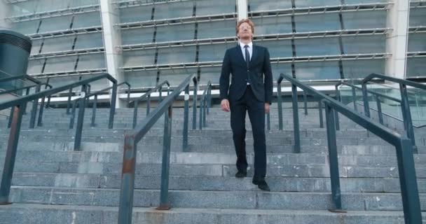 정장을 입은 빨간 머리의 젊은 사업가 가 사무실 건물에서 계단을 내려갑니다. 직장에서 좋은 하루를 보낸 남성 사업가가 계단을 걷고 있습니다. — 비디오